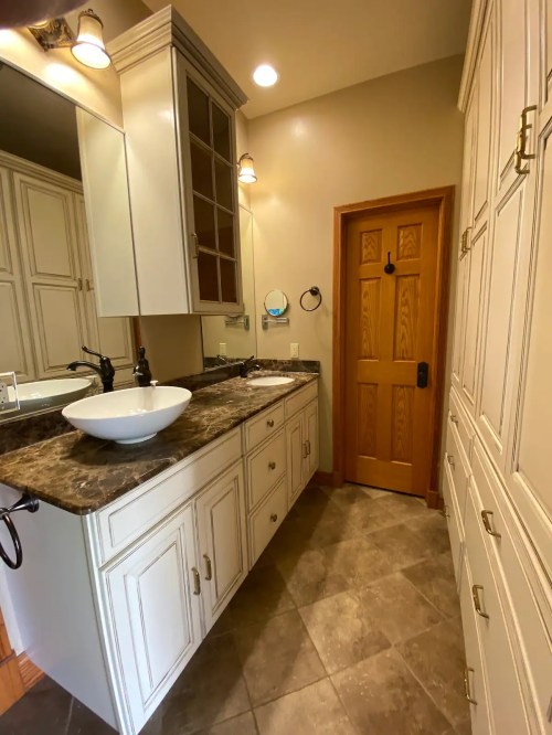 A well-lit bathroom featuring a marble countertop, a round sink, and wooden cabinetry with a door at the end.