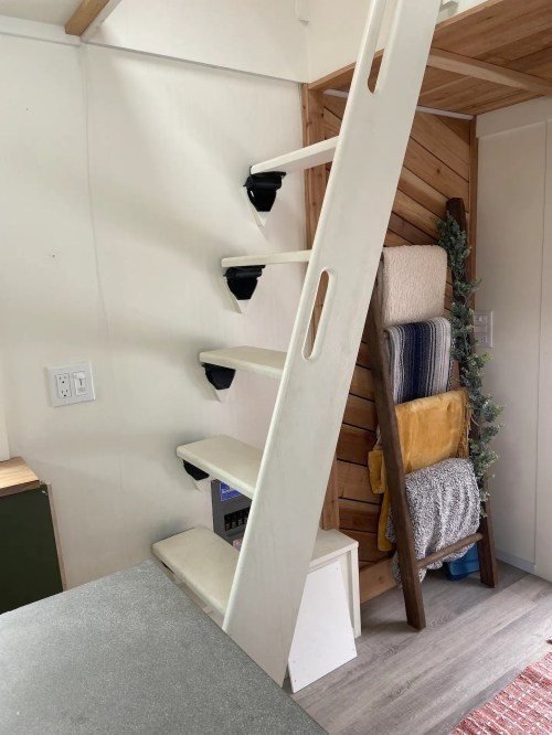 A white ladder leads to a loft in a cozy interior, with shelves displaying towels and blankets nearby.