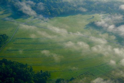 Aerial view of lush green fields with mist hovering over the landscape and a winding road cutting through.