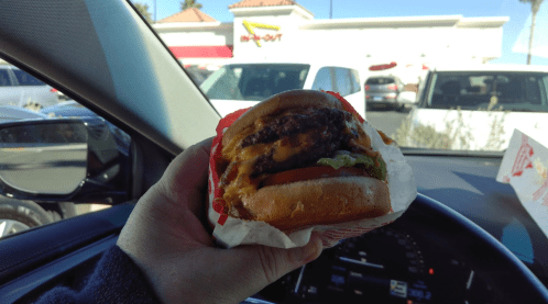 A hand holding a cheeseburger with lettuce and tomato, with an In-N-Out restaurant in the background.