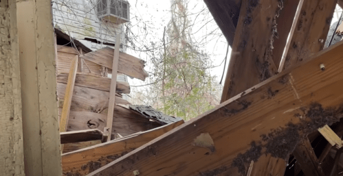 View of a damaged building interior with exposed wooden beams and debris, showing signs of decay and neglect.