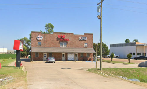 A brick building with a sign for a bar and grill, surrounded by a parking lot and greenery under a clear blue sky.