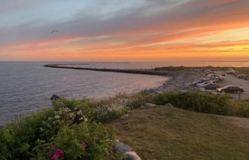 A serene coastal scene at sunset, featuring a rocky shoreline, colorful sky, and parked cars in the distance.