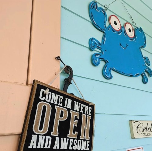 A sign reading "Come in, we're open and awesome" hangs next to a blue crab decoration on a pastel wall.