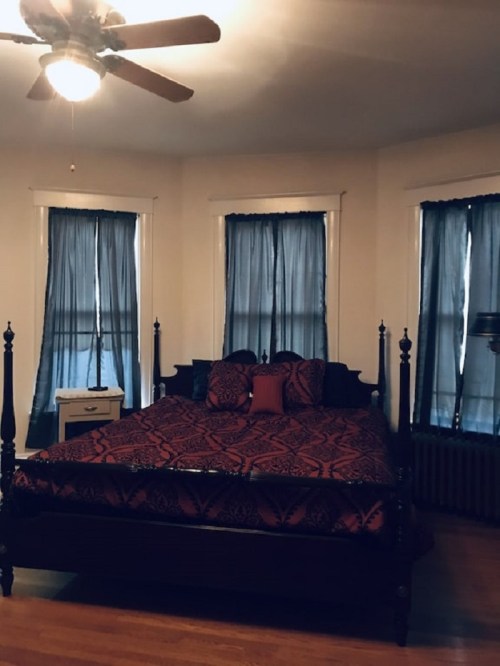 A cozy bedroom featuring a dark wood bed with a patterned red and black comforter, blue curtains, and a ceiling fan.