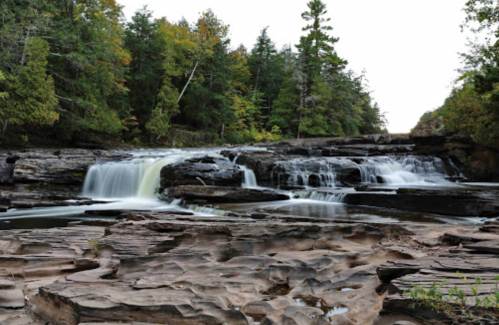 A serene waterfall cascades over rocky terrain, surrounded by lush green trees and a calm river.