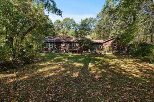 A rustic house surrounded by trees and fallen leaves, set in a serene, wooded area under a clear blue sky.