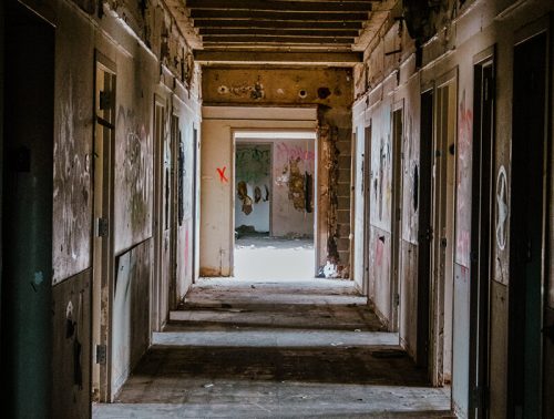 A dimly lit hallway with peeling walls and doors, featuring graffiti and light streaming from an open doorway at the end.