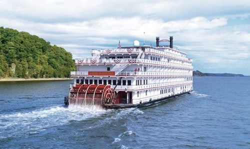 A large riverboat with paddle wheels sails on a calm waterway, surrounded by green hills under a cloudy sky.