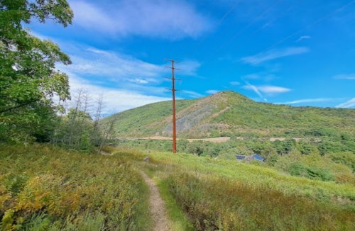 A scenic landscape featuring a dirt path, green hills, and a power line under a blue sky with scattered clouds.