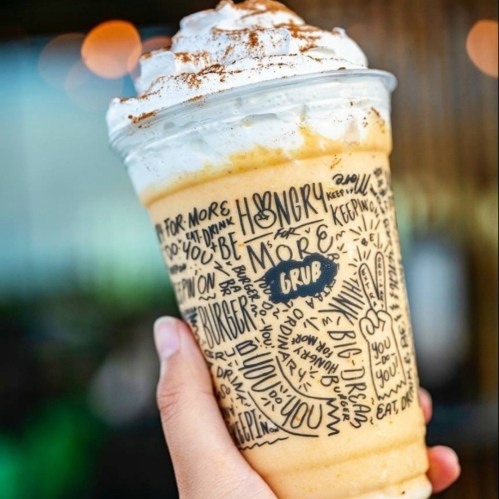 A hand holding a frosty drink topped with whipped cream and cinnamon, in a cup decorated with playful food-related words.