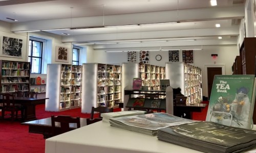 A bright library interior with bookshelves, tables, and a display of books, featuring red carpet and large windows.