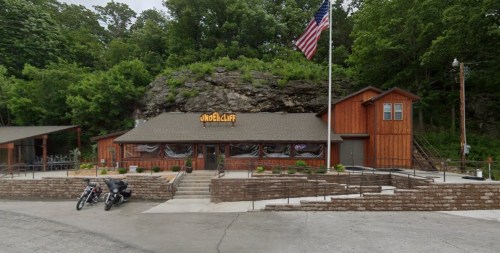 A rustic wooden building with a flag, surrounded by trees and a rock formation, featuring outdoor seating and motorcycles parked.
