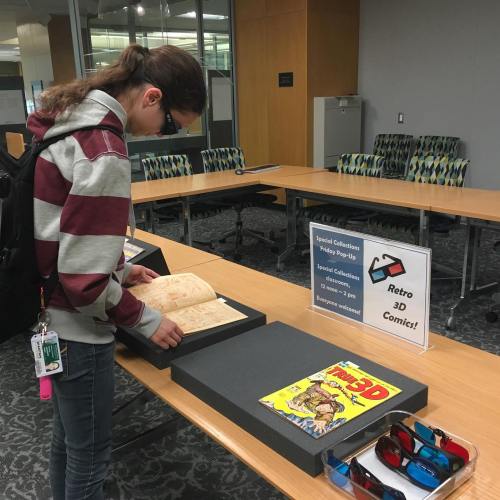 A person examines an open book at a table with a sign about a retro 3D comics event in a library setting.