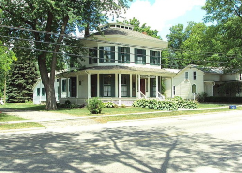 A large, two-story house with a wraparound porch, surrounded by greenery and trees on a sunny day.