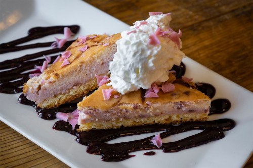 Two slices of pink dessert topped with whipped cream and pink petals, drizzled with berry sauce on a white plate.