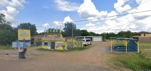 A colorful building with SpongeBob SquarePants decor, featuring a picnic area and a blue bus parked nearby under a cloudy sky.