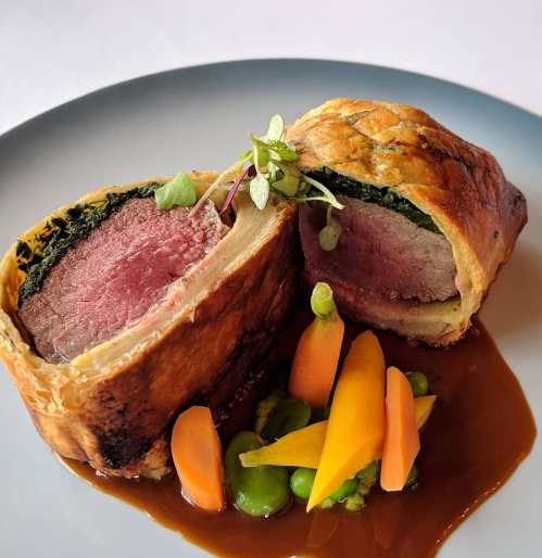 A sliced beef Wellington with spinach, served with colorful vegetables and a rich sauce on a gray plate.