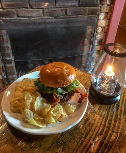 A delicious burger with lettuce and bacon served on a plate with potato chips, next to a lit lantern on a wooden table.