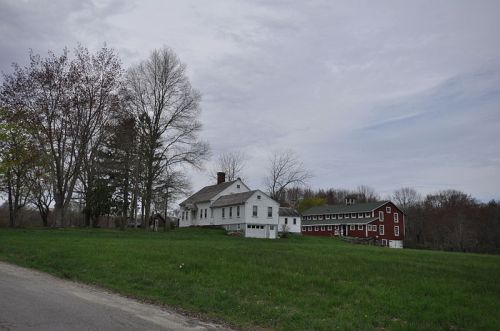 A white house and a red barn sit on a grassy landscape under a cloudy sky, surrounded by trees.