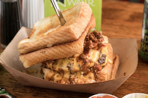 A towering sandwich with toasted bread, melted cheese, and chili, served in a paper tray on a wooden table.