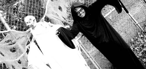 A person in a black hooded costume poses next to a white ghost figure, surrounded by cobwebs and a fence.