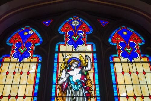 Stained glass window depicting an angel with a sword, surrounded by vibrant blue and red geometric patterns.