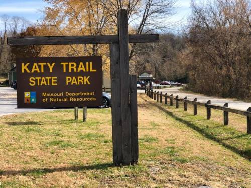 Sign for Katy Trail State Park, with trees and a gravel path in the background, indicating a natural area for recreation.