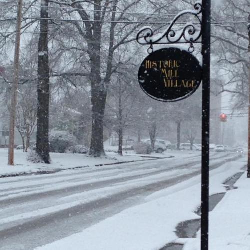 A snowy street scene featuring a sign for "Historic Mill Village" amidst falling snow and winter trees.