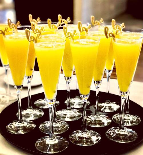 A tray of champagne flutes filled with yellow drinks, each topped with a decorative "love" stirrer.