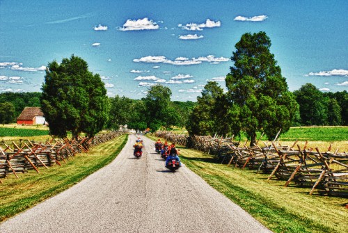 Motorcyclists ride down a scenic country road lined with trees and wooden fences under a bright blue sky.
