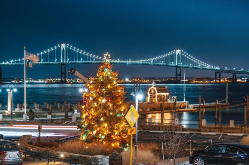 A brightly lit Christmas tree in the foreground with a bridge and city lights reflecting on the water at night.