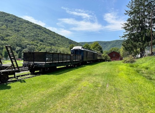 A vintage train on tracks beside a grassy area, with mountains and a red barn in the background under a clear blue sky.