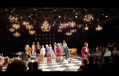 A theater performance with actors in period costumes on stage, surrounded by chandeliers and an audience.