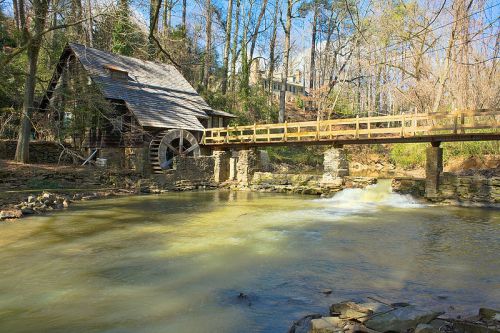 A rustic watermill beside a flowing river, with a wooden bridge and trees in the background.