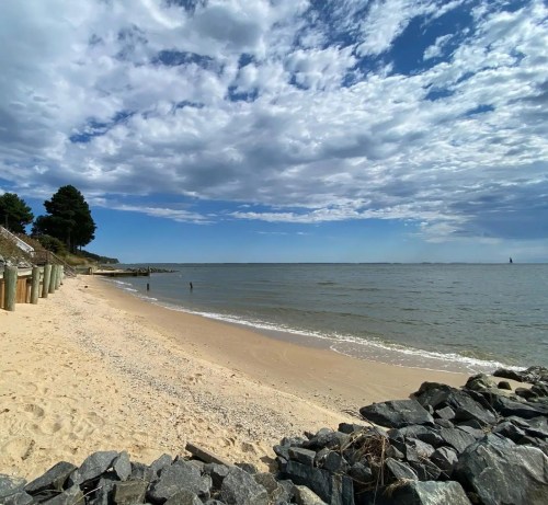 A sandy beach with gentle waves, rocky shoreline, and a cloudy blue sky in the background.