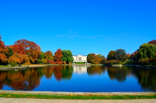 A serene park scene with a calm lake reflecting colorful autumn trees and a building in the background under a clear blue sky.