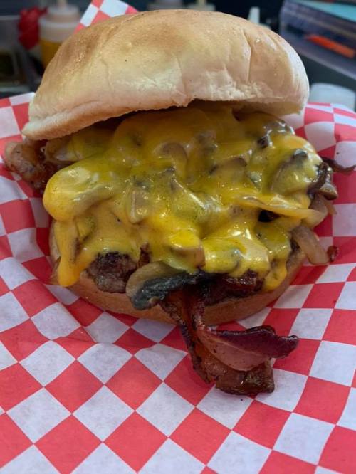 A juicy burger topped with melted cheddar cheese, bacon, and sautéed mushrooms, served in a bun with a checkered paper liner.