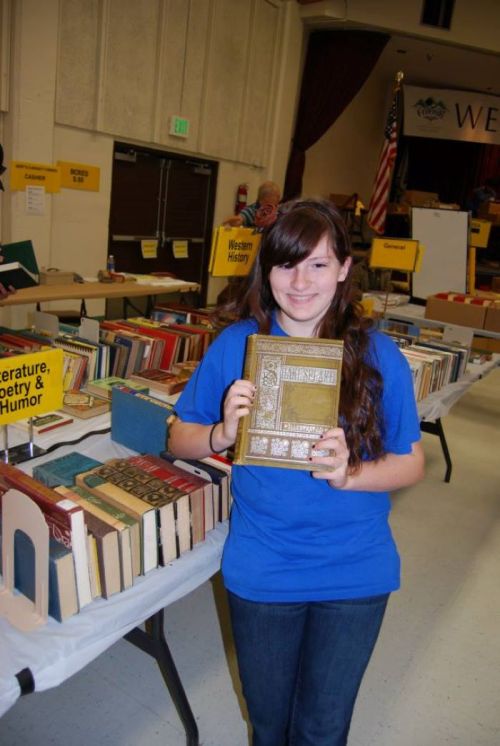A girl in a blue shirt smiles while holding a decorative book, surrounded by tables filled with various books.