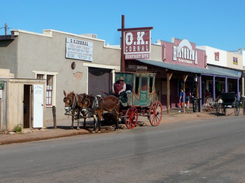 A horse-drawn carriage in front of historic buildings on a dusty street, with a sign for O.K. Corral visible.