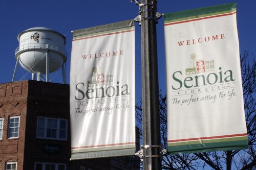 Two welcome banners for Senoia, Georgia, with a water tower in the background against a clear blue sky.