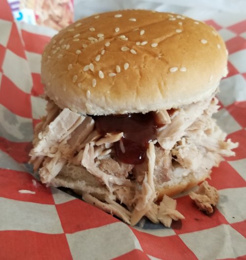 A pulled pork sandwich with barbecue sauce on a sesame seed bun, served on red and white checkered paper.
