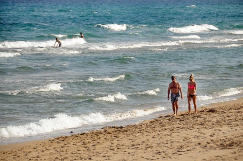 A couple walks along a sandy beach while others surf in the ocean waves under a clear blue sky.