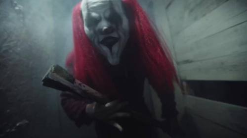 A creepy clown with red hair and a menacing expression holds a wooden stick in a dimly lit, foggy space.