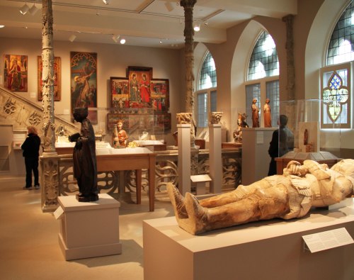 A museum interior featuring sculptures, paintings, and stained glass, with visitors exploring the exhibits.
