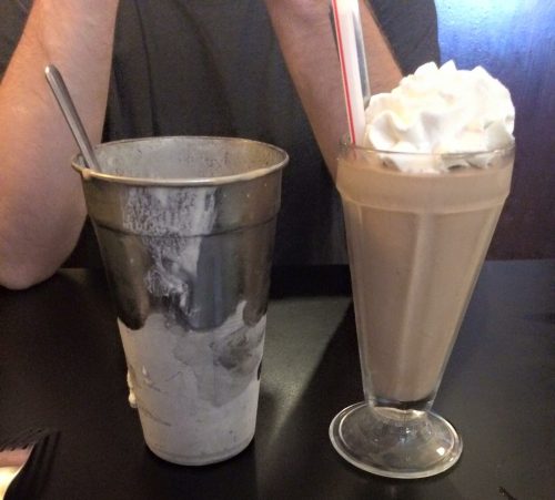 A metal cup with remnants of a milkshake next to a tall glass of a creamy milkshake topped with whipped cream.
