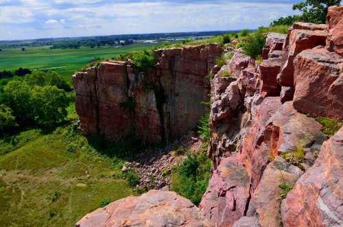 A scenic view of a rocky cliff with greenery, overlooking a vast landscape and distant fields under a blue sky.