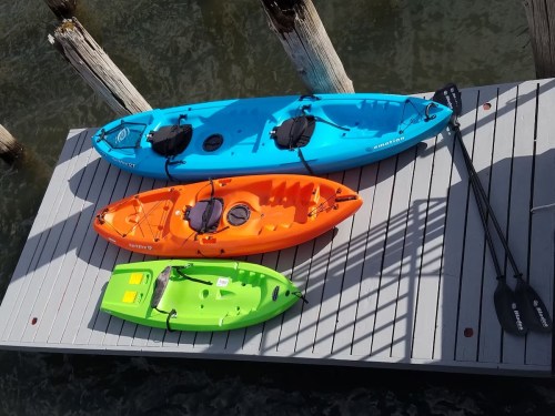 Three colorful kayaks (blue, orange, green) are lined up on a wooden dock above calm water.