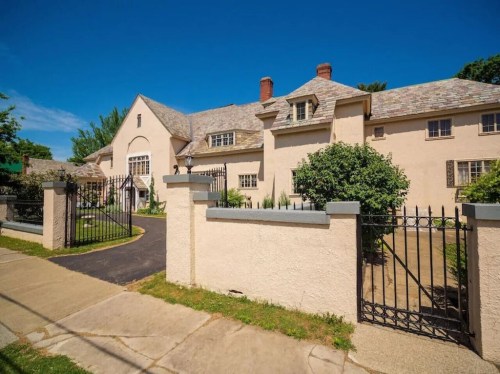 A large, light-colored house with a slate roof, surrounded by a black iron fence and green landscaping under a blue sky.