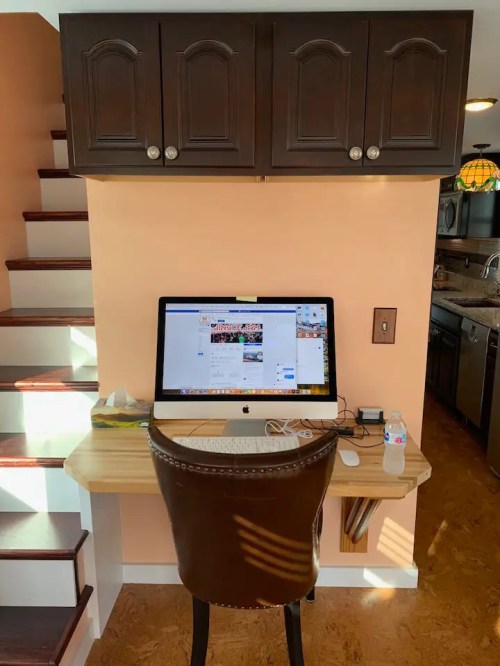 A small workspace with a computer on a wooden desk, located near stairs and kitchen cabinets, with a chair and water bottle.
